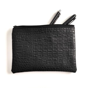 Puede incluir: Un estuche negro rectangular con cremallera y un patrón texturizado similar al de un cocodrilo. Se ven dos bolígrafos negros que sobresalen de la abertura superior. El estuche está sobre un fondo blanco.