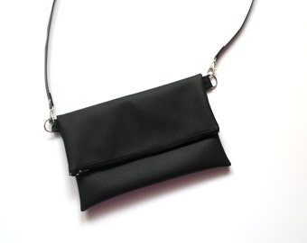 Könnte beinhalten: Eine schwarze Leder-Umhängetasche mit Klappenverschluss und Reißverschlussdetail. Die Tasche hat einen langen, verstellbaren Riemen.
