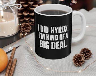 Pode incluir: Uma caneca de cerâmica preta com interior e alça brancos, apresentando o texto branco desgastado "I DID HYROX. I'M KIND OF A BIG DEAL." impresso na frente. A caneca tem um formato cilíndrico e um acabamento liso com letras claras e nítidas.
