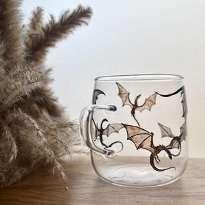 Könnte beinhalten: Ein klarer Glasbecher mit einer Reihe handbemalter brauner und schwarzer Drachenillustrationen, die um die Außenseite fliegen. Der Becher hat einen einfachen, abgerundeten Henkel und einen glatten, transparenten Körper.