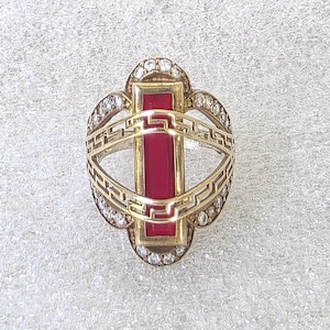 May include: A gold ring featuring a central vertical rectangular red stone. The band is decorated with a Greek key pattern and is surrounded by a border of small clear stones set in a rounded frame that encircles the central red rectangular stone.