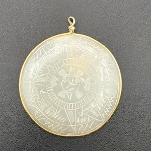 May include: A circular pendant featuring a translucent, pale green stone disc encased in a thin gold-toned wire frame. The stone surface is etched with a central sun motif surrounded by geometric patterns and radiating lines, with a small wire loop.
