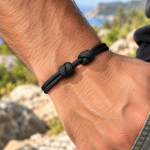 Könnte beinhalten: Ein schwarzes Armband aus zwei parallelen Kordelsträngen mit zwei markanten, fest gewebten Knoten, die mittig am Band angebracht sind. Die Kordel hat eine texturierte, geflochtene Optik und das Design ist schlicht und funktional.