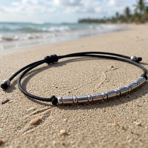 Könnte beinhalten: Ein schwarzes Kordelarmband mit einer Reihe von zylindrischen und kugelförmigen silberfarbenen Metallperlen in einem Muster. Das Armband verfügt über einen verstellbaren Schiebeknotenverschluss und zwei kleine silberfarbene Metallperlen an den Enden.