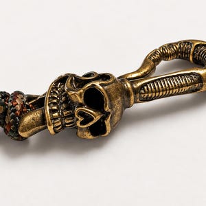 May include: A brass-toned metal swivel hook featuring a detailed skull design with a heart-shaped nose cavity. The hook is attached to a braided cord made of interwoven black, brown, and metallic copper-colored synthetic strands.