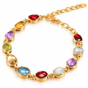 May include: A gold-tone chain bracelet featuring a series of oval-shaped, multi-coloured faceted gemstones. The stones include shades of red, yellow, purple, green, blue, and clear white. The bracelet includes a lobster claw clasp and an extender chain.