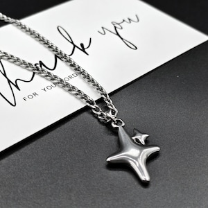 May include: A silver-toned metal necklace featuring a curb chain and a pendant shaped like a four-pointed star with a smaller star accent. The necklace rests on a white card printed with the text Thank you for your order in black script.