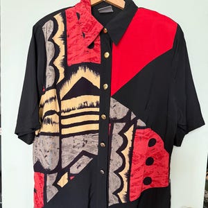 May include: A short-sleeved button-down shirt featuring a black base with a bold, abstract geometric pattern in red, tan, and gray. The shirt has a classic collar and a front placket with gold-toned buttons, showcasing a mix of solid black panels.