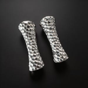 Puede incluir: Dos objetos de metal color plata con una forma cóncava similar a un reloj de arena y un acabado martillado texturizado. La superficie de cada pieza presenta un patrón repetitivo de pequeñas hendiduras reflectantes que captan la luz.