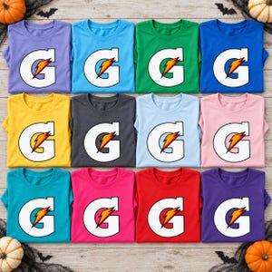 May include: A collection of twelve crew neck t-shirts in various solid colors, including purple, blue, green, yellow, gray, light blue, pink, teal, magenta, red, and dark purple. Each shirt features a large white letter G with a lightning bolt.