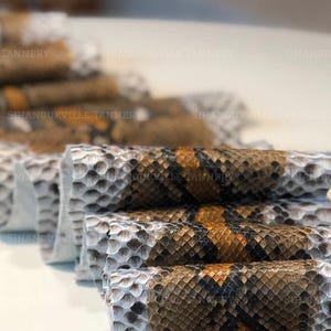 May include: A folded piece of snakeskin leather featuring a repeating pattern of white, gray, brown, and black scales. The material is arranged in a series of accordion-style pleats, showcasing the textured surface and the natural scale design.