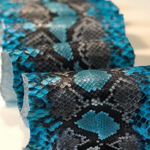 May include: A strip of snakeskin leather featuring a vibrant turquoise and black diamond-shaped scale pattern. The material is folded in a repeating wave-like formation. The text SIHANOUKVILLE TANNERY is visible across the surface.