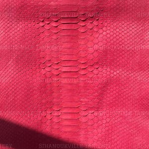 Puede incluir: Una piel de serpiente de cuero en color rosa intenso que presenta un patrón de escamas simétrico y distintivo a lo largo del centro. El material está drapeado sobre una barra horizontal, mostrando una textura uniforme y un color magenta audaz.