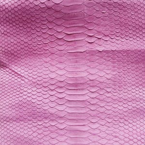 May include: A flat, rectangular piece of pink snakeskin leather featuring a distinct, repeating scale pattern. The texture consists of larger, elongated scales running down the center, flanked by smaller, rounded scales on both sides.