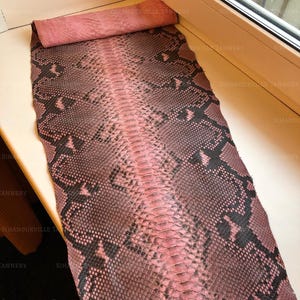 May include: A long, rectangular piece of snakeskin leather featuring a pink and dark brown scale pattern. The hide displays a distinct central ridge of smaller scales flanked by larger, diamond-shaped markings in a repeating, natural texture.