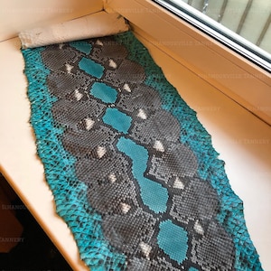 May include: A long, rectangular piece of snakeskin leather featuring a vibrant turquoise and dark gray scale pattern. The skin displays a repeating geometric design with distinct, textured scales and a natural, organic edge along the sides.