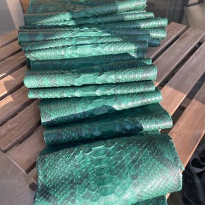 May include: A stack of emerald green snakeskin leather hides featuring a distinct, natural scale pattern. Each piece of reptile skin displays a deep, vibrant green hue with darker, subtle shading throughout the textured surface of the material.