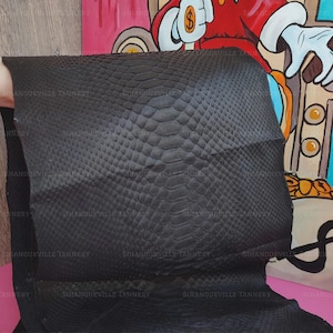 May include: A large, rectangular piece of black snakeskin leather featuring a distinct, repeating scale pattern across the entire surface. The material has a matte finish and is displayed flat, showing the natural texture and organic arrangement of the scales.