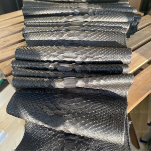 May include: A stack of folded black and silver snakeskin leather hides featuring a distinct scale pattern. Each piece of leather displays a gradient of dark charcoal to metallic silver tones. The text SIHANOUKVILLE TANNERY is visible.