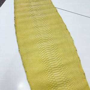 May include: A long, flat piece of yellow snakeskin leather featuring a distinct, symmetrical scale pattern that runs down the center. The material has a uniform, bright yellow color throughout, with a textured surface composed of overlapping scales.