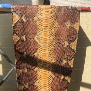 May include: A rectangular piece of snakeskin leather featuring a repeating pattern of dark brown, tan, and cream-colored scales. The skin displays a distinct central row of larger, elongated scales flanked by smaller, textured scales in a natural organic design.