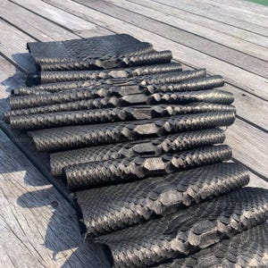 May include: A stack of multiple rectangular pieces of black snakeskin leather, each featuring a distinct, raised, and overlapping scale pattern. The material has a metallic sheen and a textured surface, arranged in a neat, layered pile on a flat surface.
