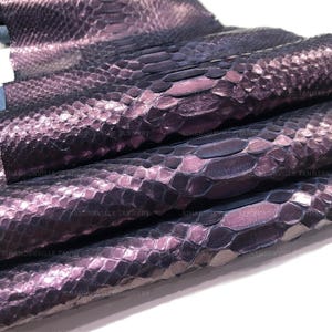 May include: A piece of snakeskin leather featuring a metallic finish with a gradient of deep purple and dark blue tones. The surface displays a repeating pattern of overlapping scales with the text SIHANOUKVILLE TANNERY visible across the material.