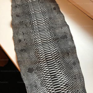 May include: A long, rectangular piece of black and white snakeskin leather featuring a distinct central row of large, elongated scales flanked by smaller, textured scales. The surface displays a repeating pattern with the text SIHANOUKVILLE TANNERY.