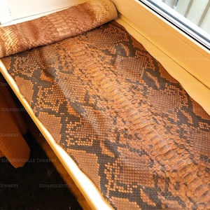 Könnte beinhalten: Ein langes, flaches Stück Schlangenleder mit einem natürlichen Schuppenmuster in Braun-, Beige- und Anthrazittönen. Das Material ist an einem Ende teilweise aufgerollt und zeigt eine strukturierte Oberfläche mit deutlichen Schuppen.