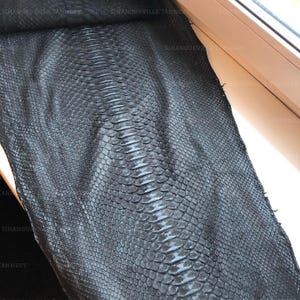 May include: A large, flat piece of black snakeskin leather featuring a distinct, symmetrical scale pattern running down the center. The surface has a subtle sheen and is marked with the repeating text SIHANOUKVILLE TANNERY across the hide.