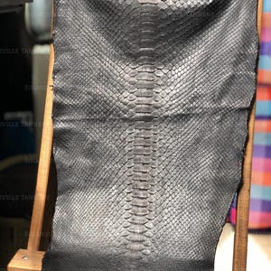 May include: A metallic charcoal gray snakeskin hide featuring a distinct, symmetrical scale pattern running down the center. The leather displays a subtle gradient effect, transitioning from a lighter, shimmering gray at the top to a deep black at the base.