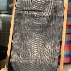 May include: A rectangular piece of dark metallic charcoal gray snakeskin leather with a distinct, repeating scale pattern. The hide is draped over a light-colored wooden frame, displaying a natural texture and a subtle sheen across the surface.