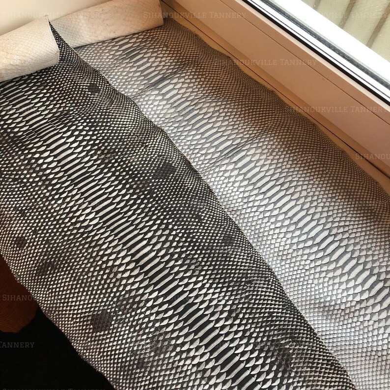 May include: Two long, rectangular pieces of textured snakeskin leather are displayed side by side. One piece features a high-contrast black and white scale pattern, while the other displays a uniform light gray scale texture throughout its length.