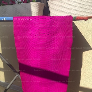 May include: A vibrant hot pink snakeskin leather hide featuring a distinct, repeating scale pattern across its entire surface. The material is draped over a horizontal bar, displaying a smooth, textured finish with a consistent, bright magenta color.