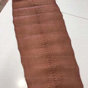 May include: A long, rectangular piece of tanned snakeskin leather in a muted brown color. The material features a distinct, repeating scale pattern that runs vertically along the center, with smaller, finer scales covering the outer edges.