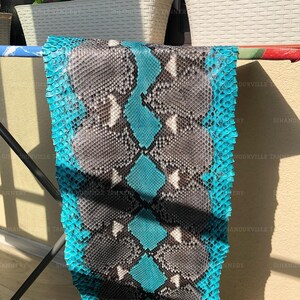 May include: A piece of genuine snakeskin leather featuring a vibrant turquoise blue background with a central pattern of large, overlapping grey and white scales. The texture is highly detailed, showing the natural scale structure of the hide.