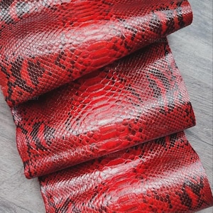 May include: A folded sheet of red and black snakeskin leather featuring a distinct reptilian scale pattern. The material displays a vibrant crimson base color with dark, irregular markings that mimic the natural texture of a snake's hide.