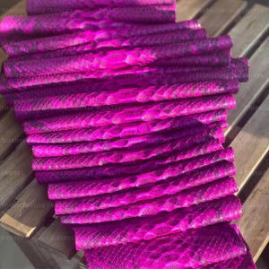 May include: A stack of multiple long, narrow strips of vibrant magenta snakeskin leather, each featuring a distinct, repeating scale pattern and a glossy finish. The text Sihanoukville Tannery is repeated across the surface of the material.