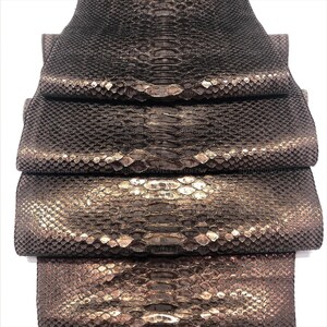 May include: A stack of four pieces of dark brown snakeskin leather with a metallic sheen. Each piece features a distinct scale pattern with a lighter, reflective center stripe running vertically down the middle of the textured hide surface.