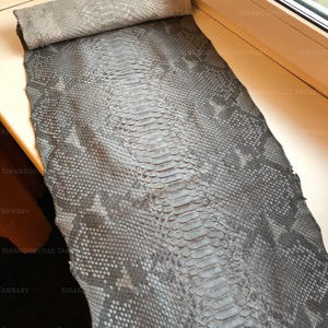 May include: A flat, rectangular piece of grey snakeskin leather featuring a natural, repeating scale pattern. The hide displays a gradient of dark and light grey tones with a textured surface, showing a central row of larger scales flanked by smaller scales.