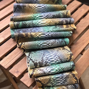 May include: A collection of rolled snakeskin pieces featuring a gradient of colors including teal, yellow, and dark brown. The scales are arranged in a repeating pattern across the surface of each roll, displaying a textured, organic appearance.