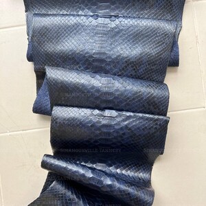 May include: A dark blue snakeskin leather hide featuring a distinct scale pattern and a glossy finish. The surface is embossed with the repeating text SIHANOUKVILLE TANNERY. The material is folded in layers, showcasing the texture and deep blue color.