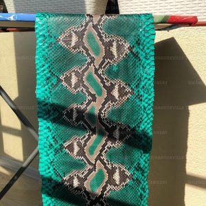 May include: A rectangular piece of emerald green snakeskin leather featuring a central vertical pattern of diamond-shaped scales in shades of black, white, and tan. The textured material displays a repeating geometric design across its surface.
