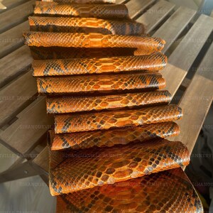 May include: A stack of multiple rectangular snakeskin leather pieces featuring a repeating pattern of orange, brown, and black scales. Each piece is folded or rolled, displaying a textured surface with the watermark Sihanoukville Tannery visible.
