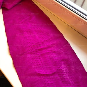 May include: A vibrant magenta snakeskin leather hide featuring a distinct, repeating scale pattern across the entire surface. The material displays a rich, saturated pink hue with a textured finish and the text SIHANOUKVILLE TANNERY repeated.