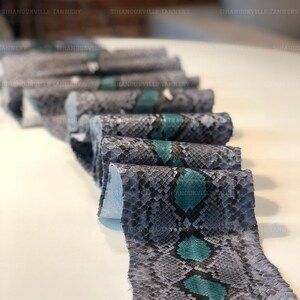 May include: A long, continuous strip of snakeskin leather featuring a repeating diamond pattern in shades of grey, black, and vibrant teal. The material is folded in multiple layers, displaying the textured, scaled surface and the text SIHANOUKVILLE TANNERY.