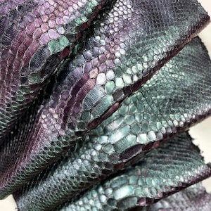 May include: A stack of layered leather sheets featuring a textured snakeskin pattern with an iridescent finish. The material displays a gradient of deep purple, metallic green, and dark charcoal tones across the overlapping, scale-like surface.