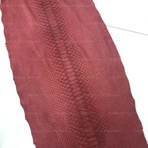 May include: A deep burgundy snakeskin hide featuring a prominent central row of large, elongated scales flanked by smaller, uniform diamond-shaped scales across the entire surface, displaying a consistent matte texture and rich, dark red coloration.