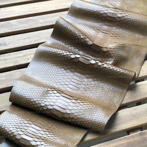 May include: A tan-colored snakeskin leather hide featuring a distinct, overlapping scale pattern across its entire surface. The material is folded in several sections, displaying a natural, textured finish with the text SIHANOUKVILLE TANNERY.