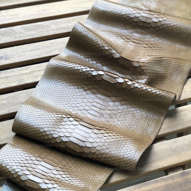 K&ouml;nnte beinhalten: Ein lohfarbenes Schlangenleder mit einem ausgepr&auml;gten, &uuml;berlappenden Schuppenmuster auf der gesamten Oberfl&auml;che. Das Material ist in mehrere Abschnitte gefaltet und zeigt eine nat&uuml;rliche, strukturierte Oberfl&auml;che mit dem Text SIHANOUKVILLE TANNERY.